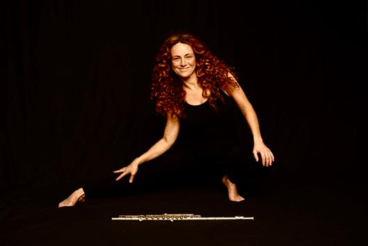 Woman in a black bodysuit against a black background doing a lunge with a flute lying on the ground in front of her.